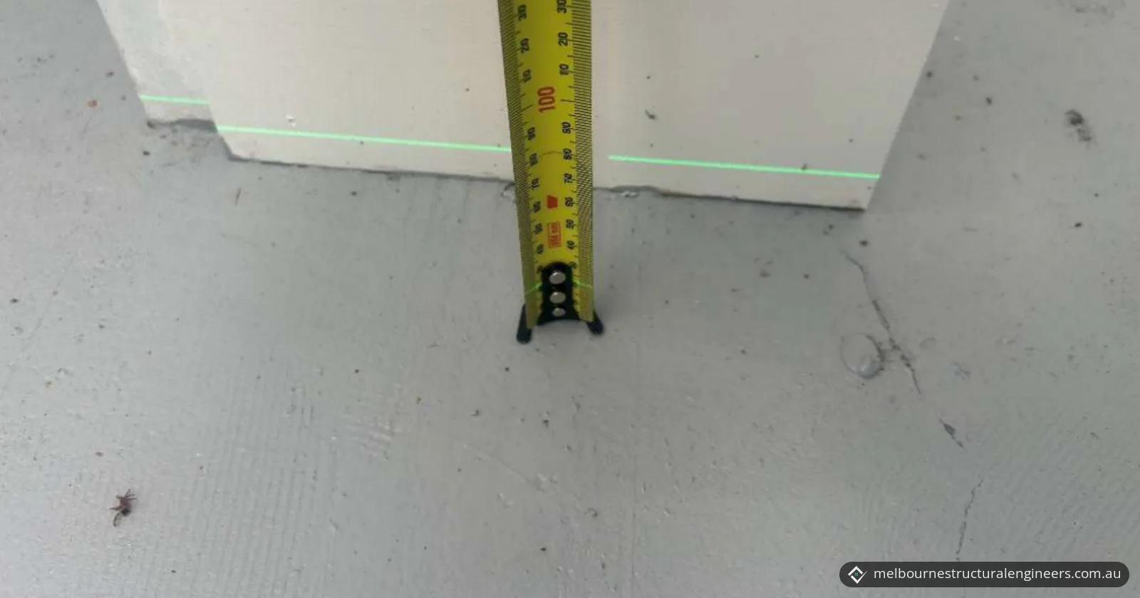 A tape measure standing on a concrete floor against a wall, with a green laser level line across the wall