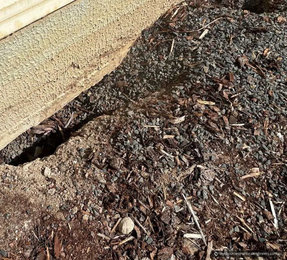 A gap opening under the rendered base of an external wall, beside gravel and mulch