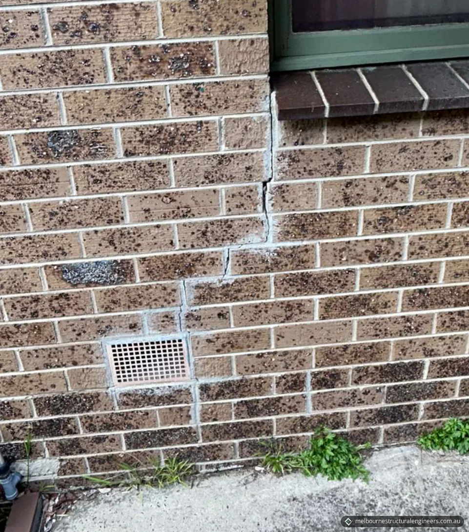 Stepped crack running down face brickwork from the corner of a window sill past a wall vent